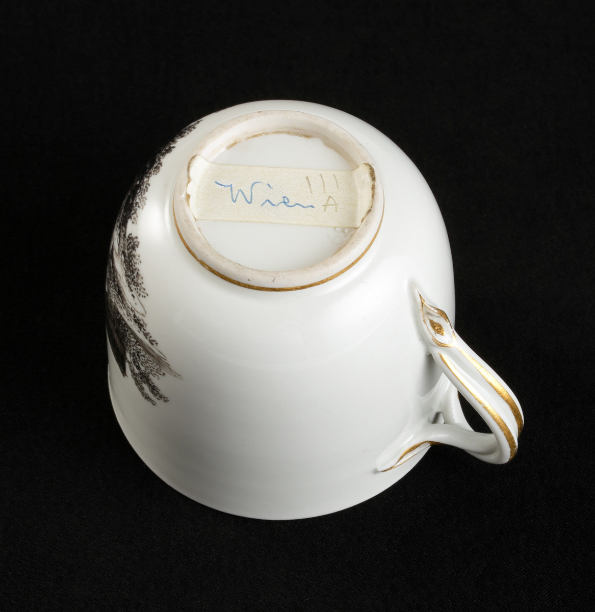 Kopp med fat av porslin. Koppen i bägarform. Fatet skålformigt. 
Dekorerad med ruinlandskap i svart camaïeu, med färglagda figurer. 
Kring kanterna spetsliknande bårder i guld. 

Inskrivet i huvudbok 1967.