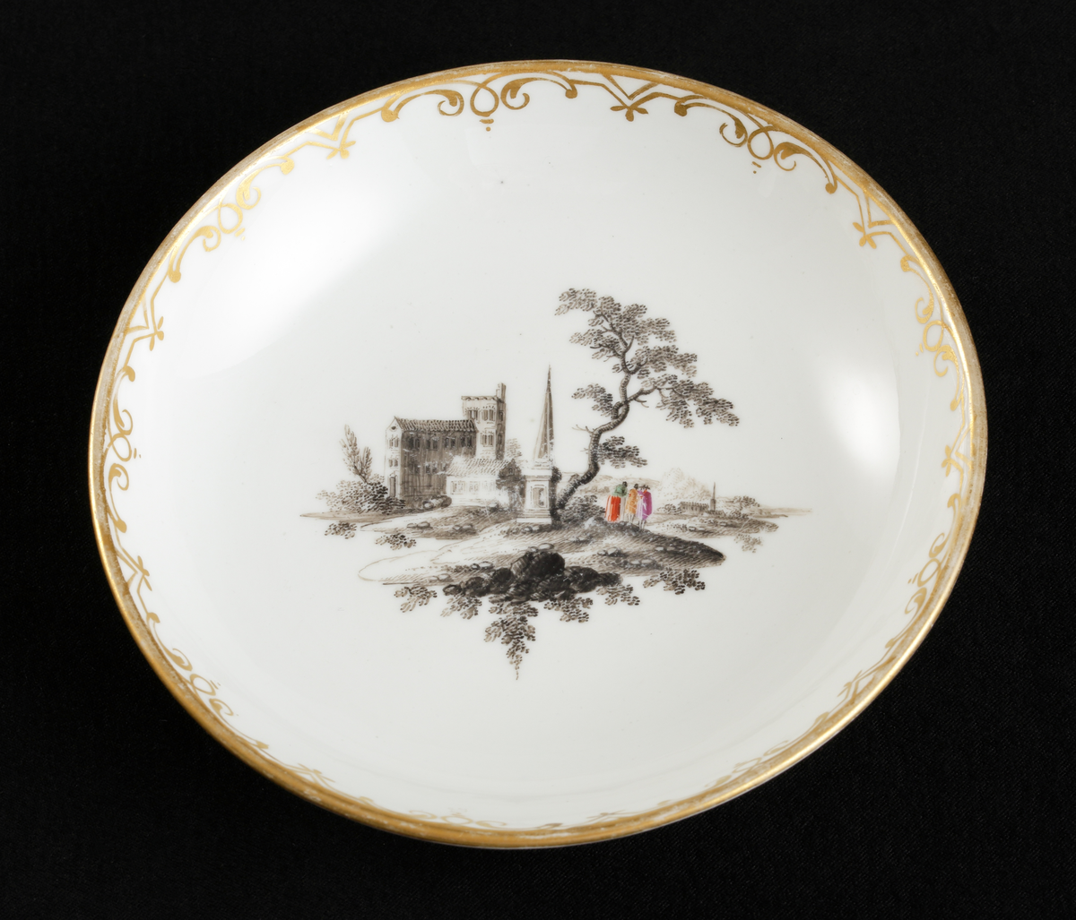 Kopp med fat av porslin. Koppen i bägarform. Fatet skålformigt. 
Dekorerad med ruinlandskap i svart camaïeu, med färglagda figurer. 
Kring kanterna spetsliknande bårder i guld. 

Inskrivet i huvudbok 1967.