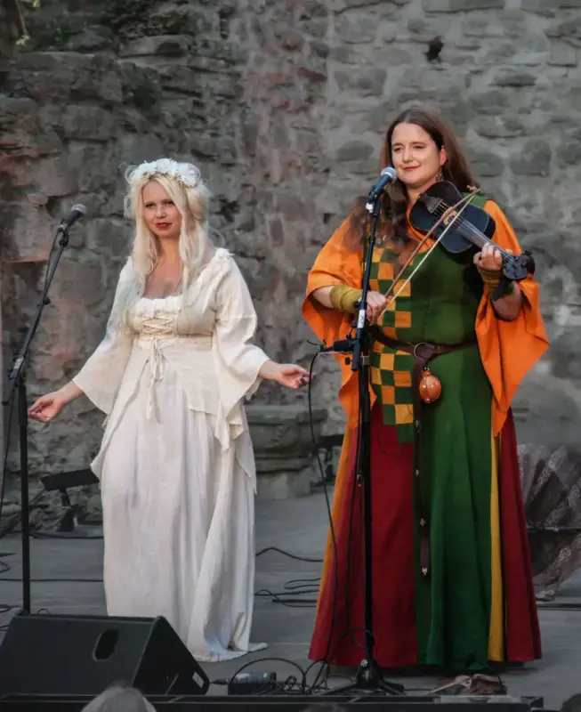 Kvinne i hvit middelalderdrakt med hvite blomster i håret som synger. Kvinne i oransje, gul, grønn og rød middelalderdrakt som spiller fiddle/middelalderfele