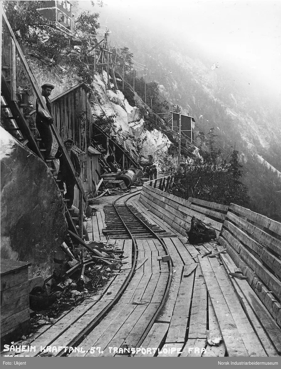 Smalsporet jernbane for transport av materialer og masse til fordelingsbassenget for Såheim Kraftstasjon. Brakker i fjellsiden. Arbeidere stående i trapp.