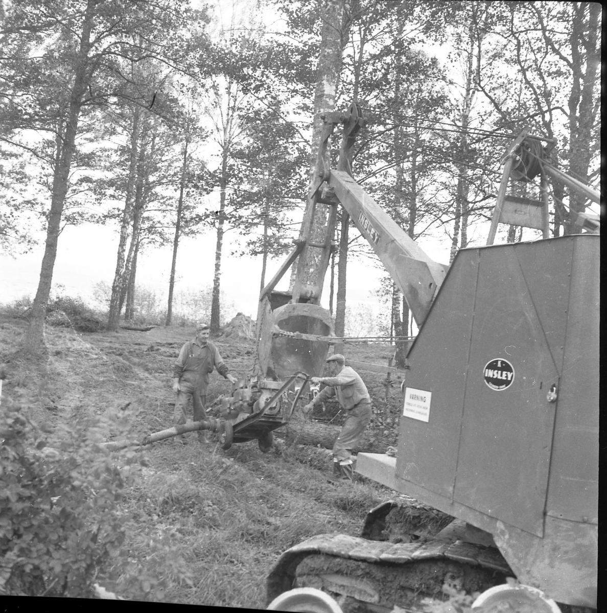 En grävmaskin gräver hål för vatten- och aloppsrör. Två män står och arbetar vid skopan.