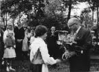 En tunnhårig man med glasögon tar emot en blomsterbukett av en flicka i folkdräkt. Mellan dem, i bakgrunden, applåderar en man. Folksamling runtomkring.