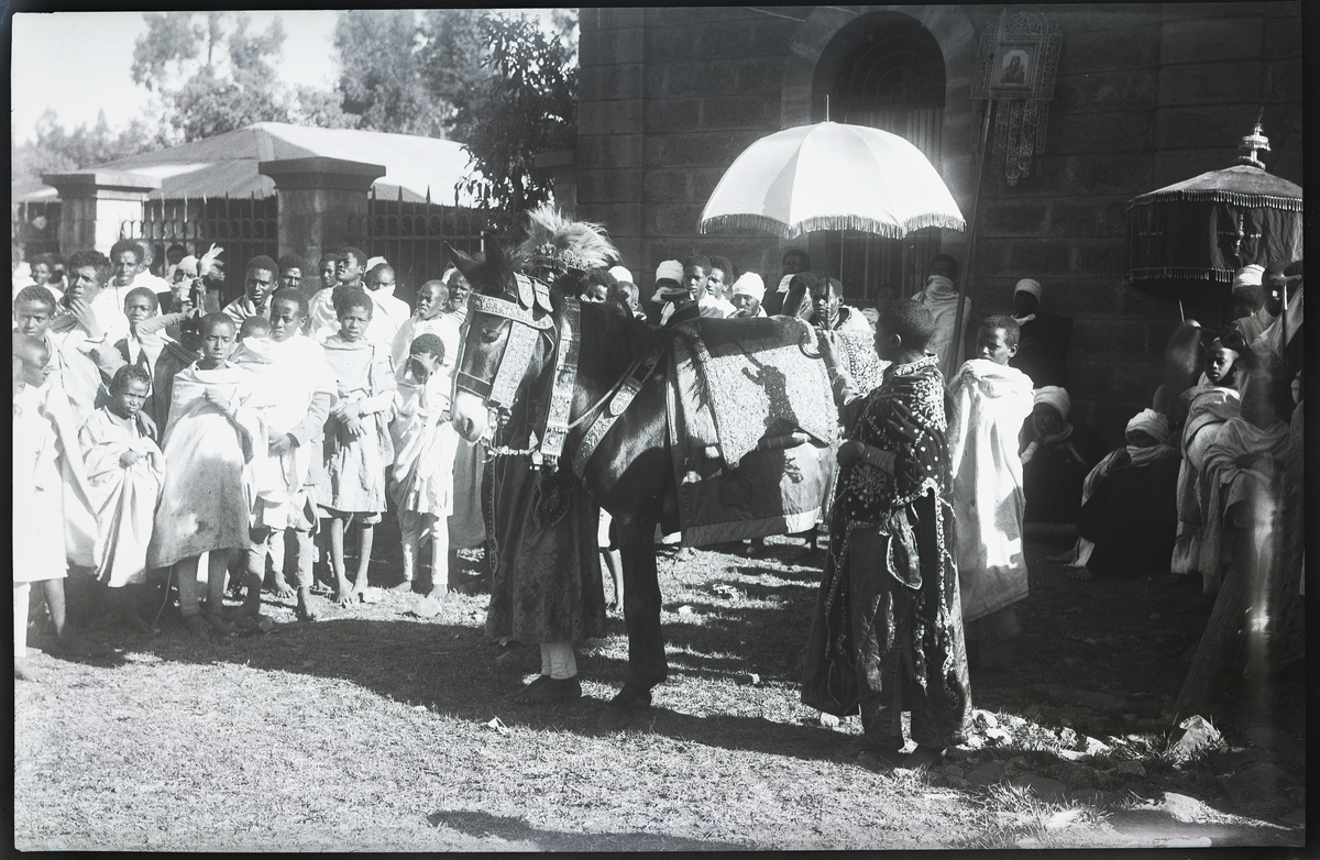 Foto tatt under reise i Afrika. Folkemengde rundt marked i Addis Ababa, hovedstaden i Etiopia 

Dekorert og pyntet esel med to velkledde personer på hver sin side. 

Påskrift på konvolutt negativ lå i: Adis Ababa