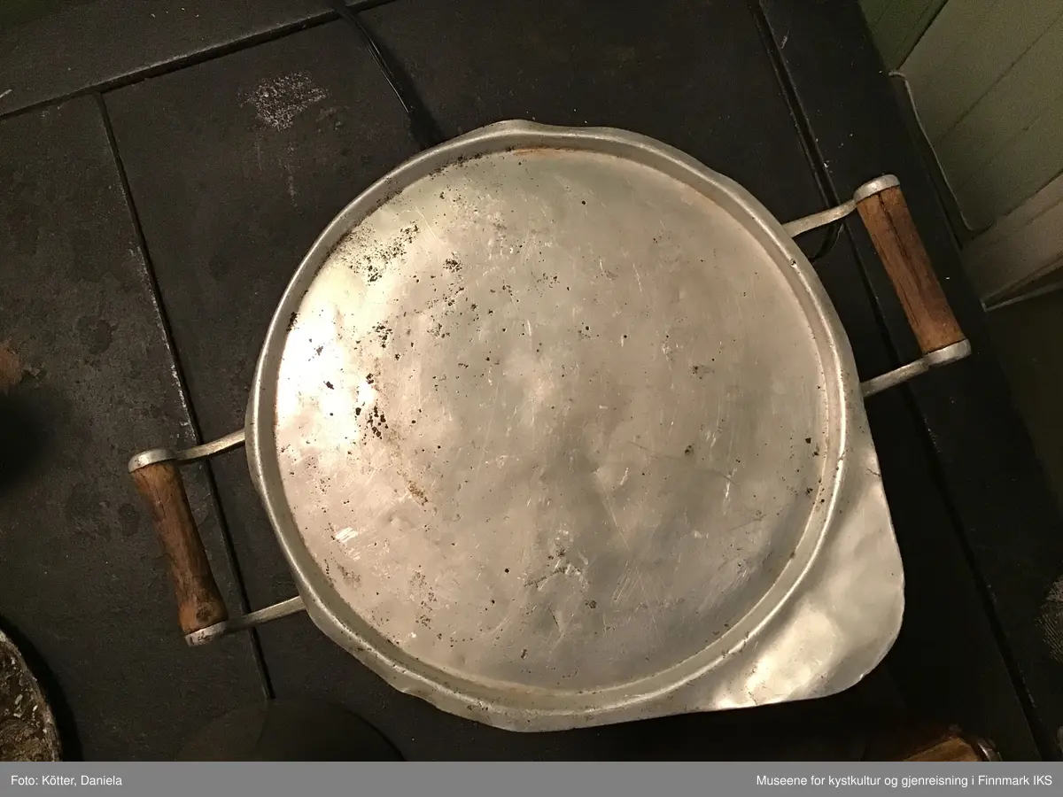 Rund, flat aluminiumslokk med kant og et grep i form av et avrundet hjørne.