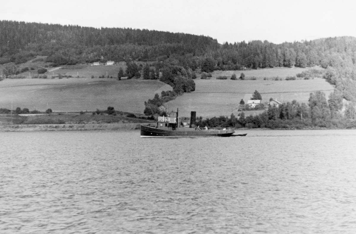 Slepebåt på Mjøsa, muligens slepebåten «Thor», som var bygd for Norsk Hovedjernbane i 1875, men som etter å ha blitt overtatt av andre eiere omkring 1900 hovedsakelig gikk som tømmersleper fram til 1954. Den avbildete båte var malt i mørke farger, med unntak av et rekkverk rundt brua foran styrhuset, som var kvitmalt. Bak det dampdrevne fartøyet hang det en liten robåt på slep. Den ble ventelig brukt det skulle ordnes med slepene.  I bakgrunnen ser vi ei li ned mot innsjøen. I den nedre delen av lia gikk det veg- og jernbanetraseer langs stranda med kornåkrer ovenfor. Høyere oppe mot åskammen sto skogen tett. Landskapet minner om Ringsfjerdingen i Ringsaker prestegjeld.