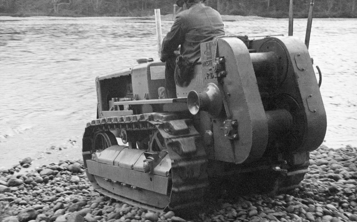 Beltekjøretøy med bakmontert vinsj, brukt under «elverensingsarbeid» i elva Flisa i 1938. Kjøretøyet et fotografert bakfra på ei steinete strand. Maskinføreren er iført vindjakke og hatt.  Han sitter med ryggen til fotografen. I bakgrunnen ses et bredt og forholdsvis stilleflytende parti av Flisa.