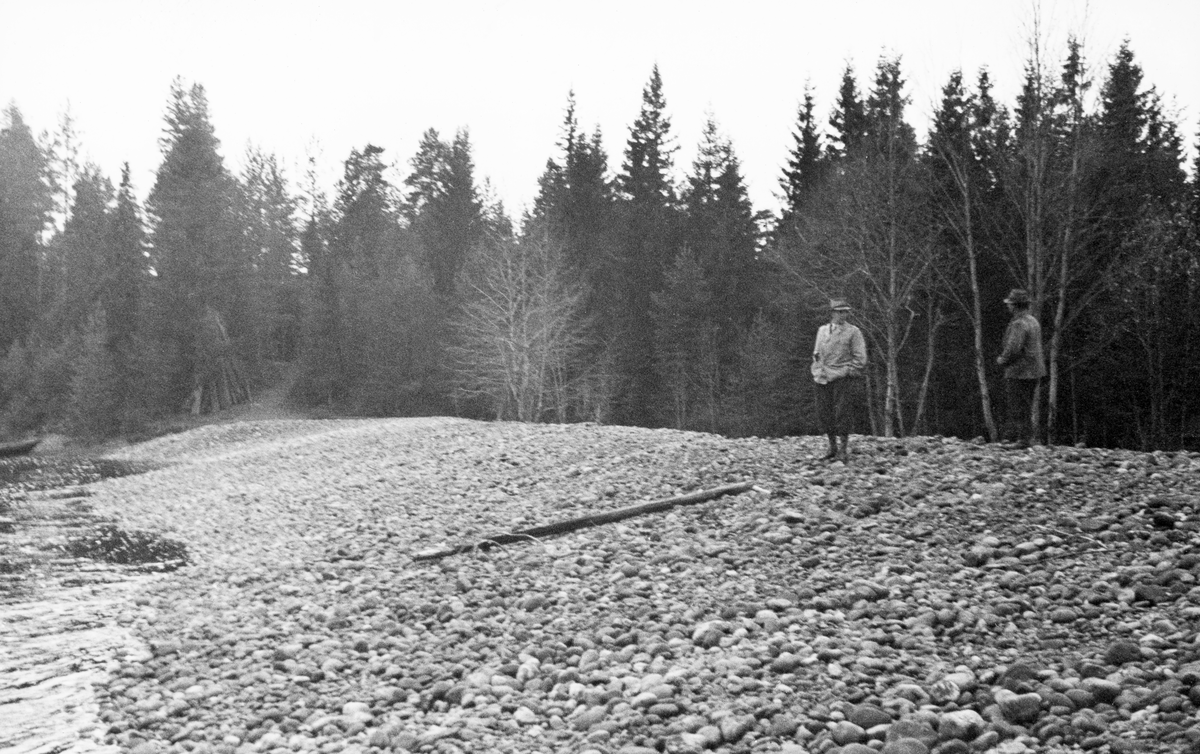 Fra opprenskingsarbeider i elva Flisa i 1938.  Fotografiet er tatt mot og langsetter en rygg med halvstor stein langs elvebredden.  På denne ryggen står fløtingsinspektøren, Kaare Sparby, og vinsjføreren.  Foran dem ligger det en stokk på elvebakken.  Bakenfor vokser det enkelte lauvtrær (antakelig or) foran et belte av granskog. 