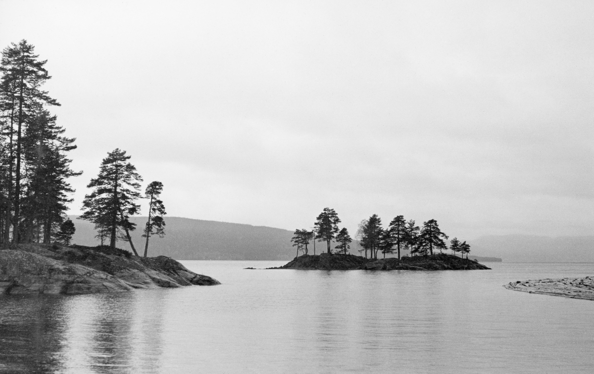 Fra Hurdalssjøen i Akershus våren 1939. Fotografiet er tatt ut over det blanke vannspeilet med en berglendt odde til venstre og en liten holme sentralt i bildet. Både på odden og på holmen vokste det furutrær. Helt til høyre i bildet skimter vi en liten del av en tømmerbom.