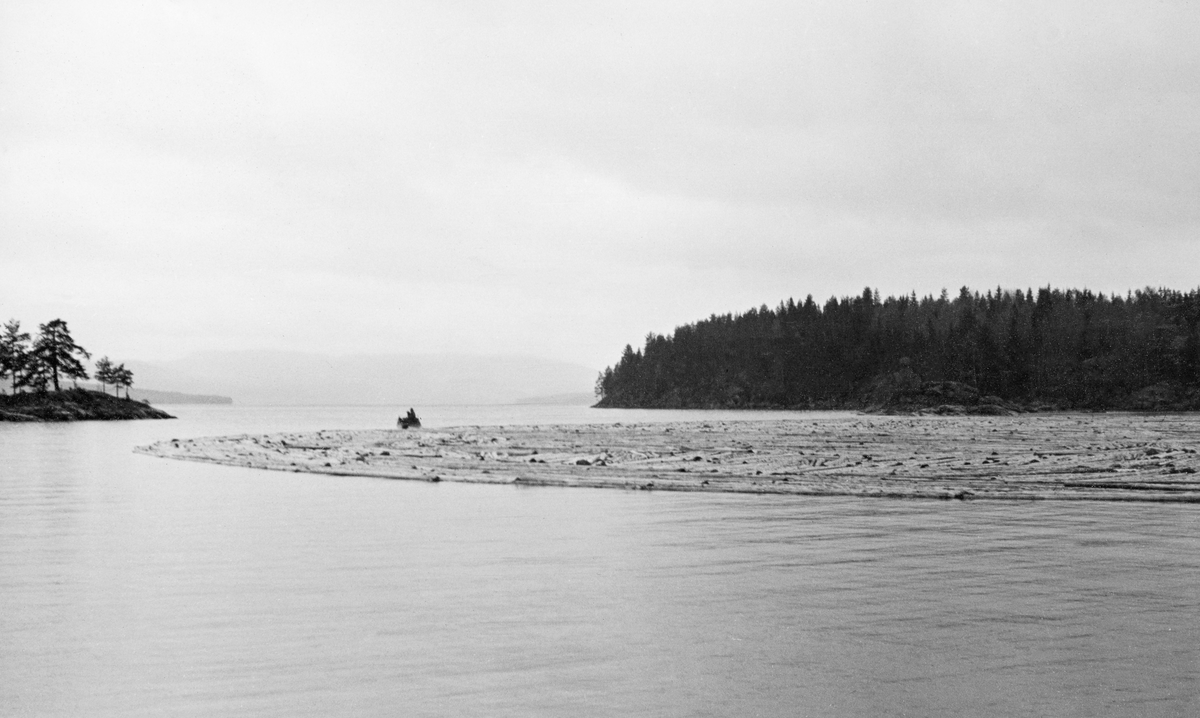 Fra Hurdalssjøen i Akershus våren 1939. Fotografiet er tatt ut over det blanke vannspeilet der det lå en ringbom med tømmer. På den andre sida av bommen skimter vi en liten båt. På frunn av avstanden fra stranda bildet er tatt fra er det usikkert om dette var en varpebåt eller en robåt.