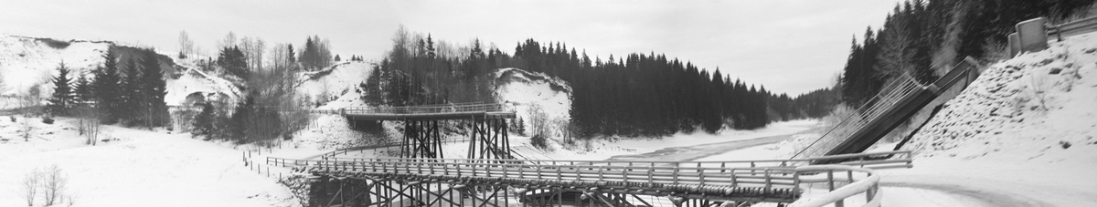 Den 18. april 1940 meldte avisa Øvre Smaalenene Dagblad at Mønster bru over Østelva i Tosebygda i Trøgstad kommune var sprengt. Brua lå noen få hundre meter fra elvas utløp i den søndre enden av innsjøen Øyeren. På dette fotografiet, som antakelig er tatt cirka et halvår etter denne hendelsen, seinhøstes i 1940, ser vi den ødelagte brua i bakgrunnen og ei nyoppbygd, noe lavere bru foran. Til erstatningsbrua var det bygd en ny vegtrasé litt lengre nede i elveskråningen enn den vegen som gikk til den høye brua. Fotografiet er tatt etter at det hadde falt snø over landskapet og etter at det var dannet ei isskorpe på elva. Under den tyske invasjonen i april hadde norske militære plassert sprengstoff under Mønster-brua, og ifølge avisa var dette blitt detonert ved en «feiltagelse». «Noen stor militær betydning kan denne bru ikke sies å ha, og i særdeleshet ikke slik som situasjonen har utviklet sig. Halvparten av brua skal være skutt vekk», rapporterte avisa. Det ble forholdsvis raskt lagt ei klopp over vassdraget der gående og syklende enkelt kunne passere. Høsten 1940 bevilget fylkesvegkontoret i Østfold 20 000 kroner til gjenoppbygging av Mønster-brua i Trøgstad. Arbeidet ble likevel ikke igangsatt umiddelbart fordi man regnet med å få lettere tak i jernbjelkene på elvebotnen når vannstanden sank litt. Sannsynligvis er det erstatningsbrua vi ser bak den istykkersprengte på dette fotografiet. Dette er et «vidvinkelopptak», sammensatt av to bilder som er tatt med normalobjektiv.