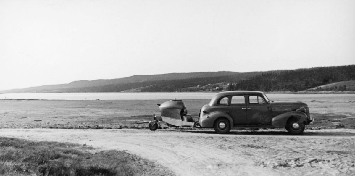 Fra fløtinga i Osensjøen i 1941. Hovedmotivet her er en bil, antakelig fløtingsinspektørens bil, med et etterhengt hjulgående apparat, muligens en trekolgenerator. Bilen - antakelig fløtingsdirektør Johs Johannesens 1937-modell Studebaker - er fotografert parkert på vegen ved sørenden av Osensjøen.  I bakgrunnen ses sjøen med bakenforliggende åser med gardsbruk i hellingene ned mot sjøen.