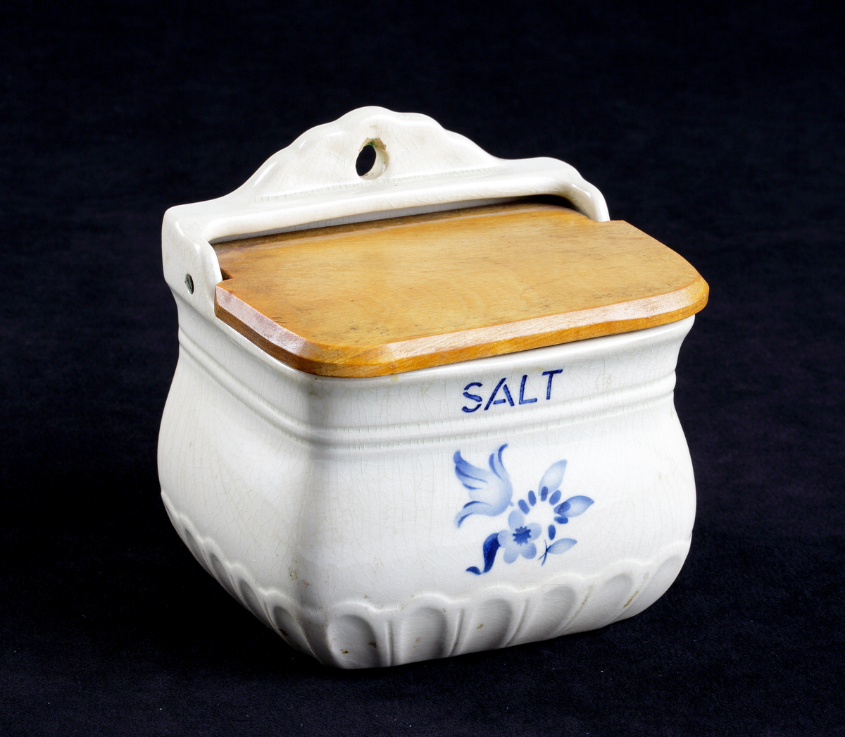 Burk av vitt porslin, med fällbart lock av fernissat trä. På framsidan dekor i form av ett blommotiv i blått, 
samt däröver ordet "Salt".

Inskrivet i huvudkatalog 1983.