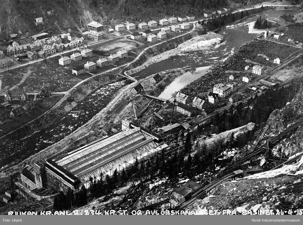Rjukan sentrum med boligbebyggelse og næringsbygg, sportsplassen og Rjukan torg. Bebyggelse på Granli og Mælandstangen. Såheim kraftstasjon under bygging. Anleggsbrakker og anleggsveier i fjellsiden over Såheim. Sett fra fordelingsbassenget.