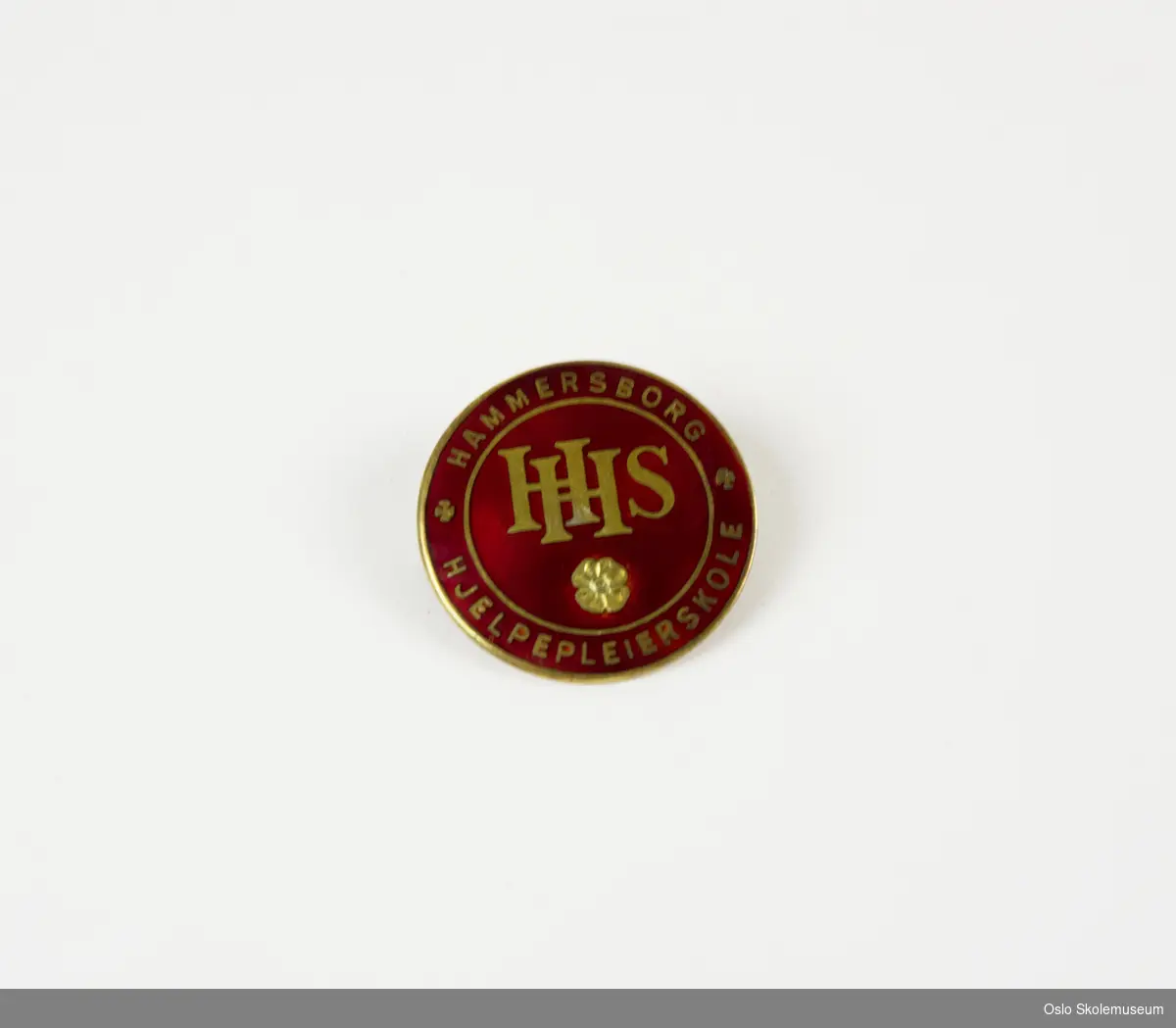 Rundt merke i metall. På fremsiden er det avbildet bokstavene "HHS" samt en blomst i gull på rød bakgrunn. Rundt stjernen står teksten "Hammersborg - Hjelpepleierskole". På baksiden er det en nål for å kunne feste merket til klær.