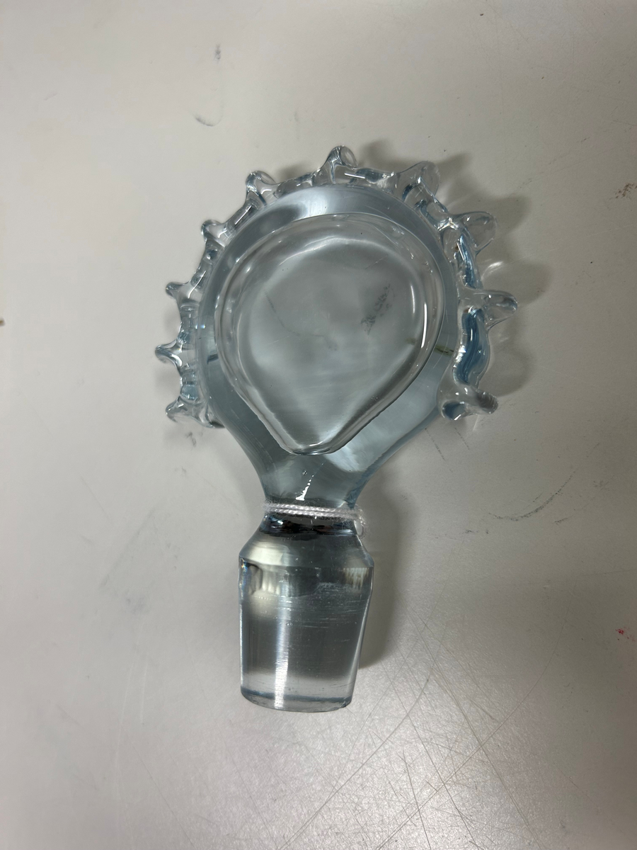 Karaff med propp, av blåtonat glas. Rund fot, flat form, intryckt på mitten, kort hals med utvikt mynningskant, strålformad dekor på ytterkanter och propp.