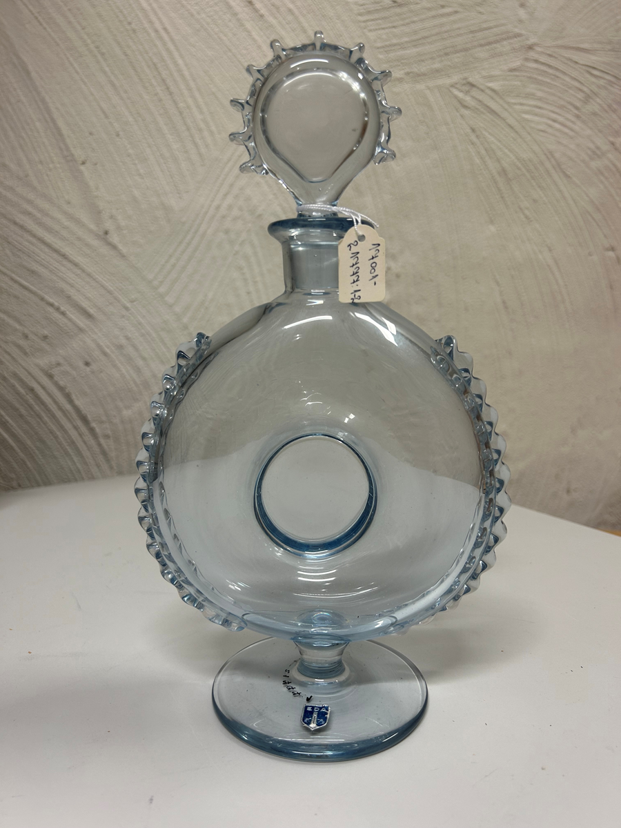 Karaff med propp, av blåtonat glas. Rund fot, flat form, intryckt på mitten, kort hals med utvikt mynningskant, strålformad dekor på ytterkanter och propp.