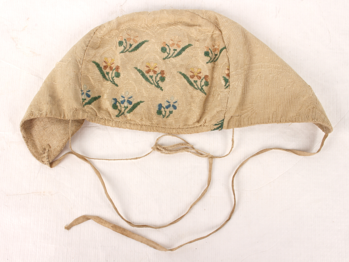 Hovedstoff beinhvit silke, ripsvev med innvevd mønster (blomsterranker + bølgemønster og prikker). Noen steder har blomstermønsteret farger. Fôr i grovt ubleika linlerret. Smale knytebånd. Snor til stramming bak. 
Pullen har tett blomstermønster. Pannestykket har et åpnere mønster, men helt i samme stil. Sannsynligvis omsøm fra samme gamle plagg. Lite innskjøtt stykke midt på pannestykke øverst.