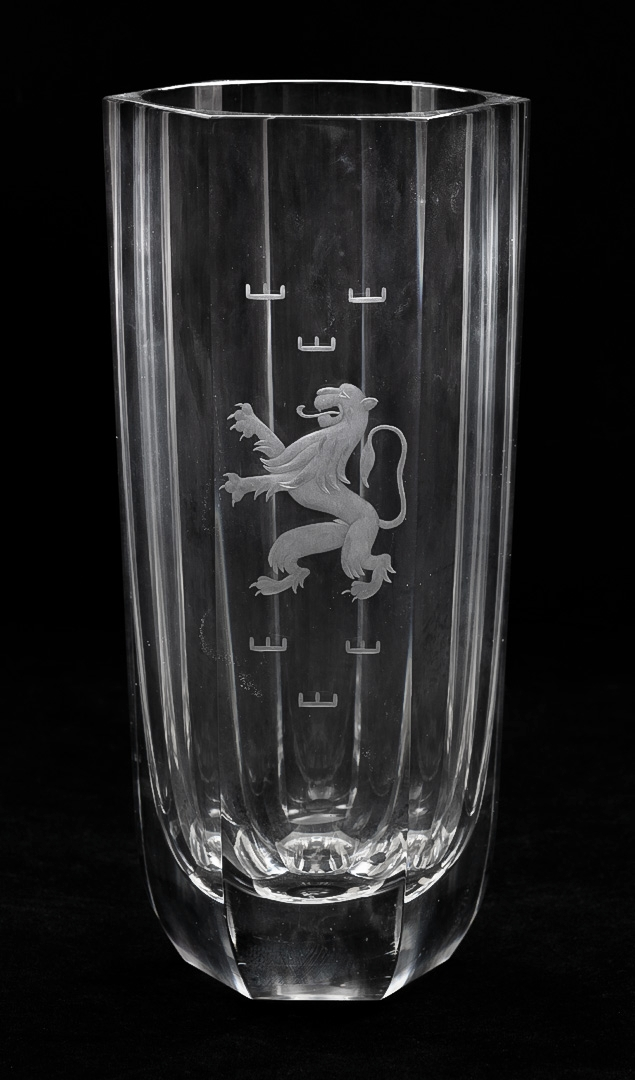 Høyreist vase i klart glass, dekorert med gravering. Fasettert heksagonal ytterside, samt sirkulær innside. På forsiden er den svenske løven flankert av to frtemstillinger av de tre kroner. Tykk planslipt bunn.