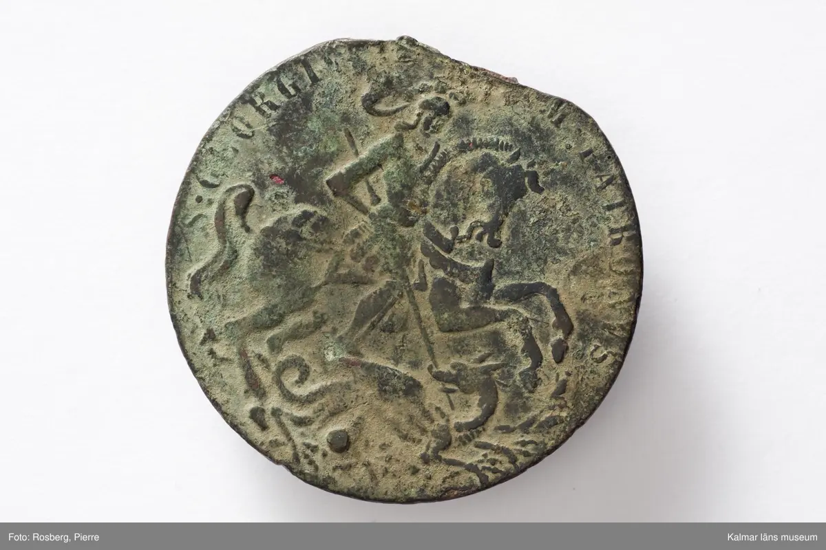 KLM 44234. Medaljong, skyddsmedaljong. Av brons. Sankt Göran-medaljong. Vikt 17,87 g. Avbrutet hänge. Åtsidan, motiv: S:t Göran och draken, text: S:GEORGIVS EQVITVM PATRONVS (S:t Georg, ryttarnas skyddspatron). Frånsidan, motiv: Kristus och hans lärjungar ombord på ett segelfartyg under storm, text: IN TEMPESTATE SECVRITAS (säkerhet i stormen).