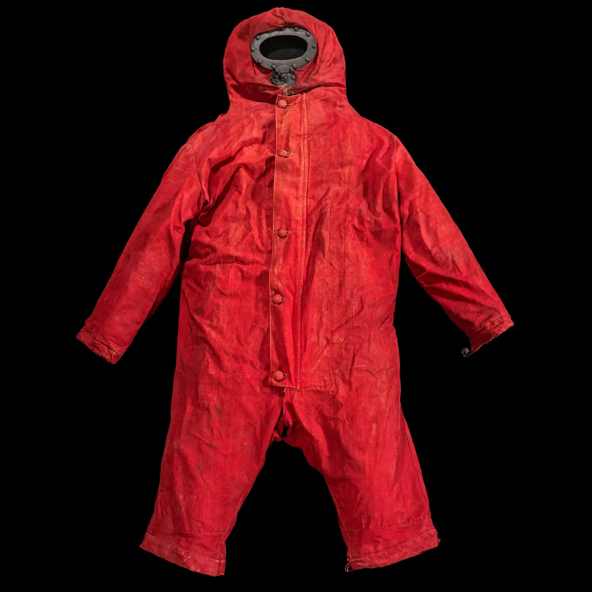 Dräkt av rött linne. Täcker huvud, armar, lår och ben till knät. Knäpps framtill med runda knappar klädda i samma textil som resterande av dräkten. Innertyget är ofärgat och knäpps med stora runda svarta knappar med ett rutmönster. Gjorda av hård plast. Insidan av det inre tyget är täckt med ett gummiliknande material, troligen guttaperka.  På huvudet, vid bärarens ansikte, är en järnrem infattad med oval glasskiva framtill. Nedanför glaset är ett hål (även detta omringad av en järnrem) som går att täppa igen med en rund korkbit. Korkbitens ena sida är täck med järn och en ögla i mitten där en kedja är fast i järnbården runt hålet. 
