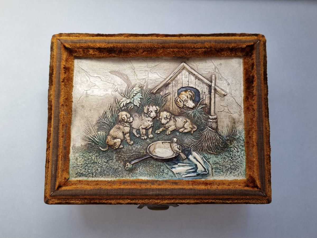 Syskrin i brun sammet. Locket är dekorerat med pressat läder (läderpressning) med motivet: Fyra hundar, en fågel och hundkoja. Invändigt vadderat med brun satin. Skrinet innehåller sax och nåletui i ben (?). Två mässingsfötter saknas.