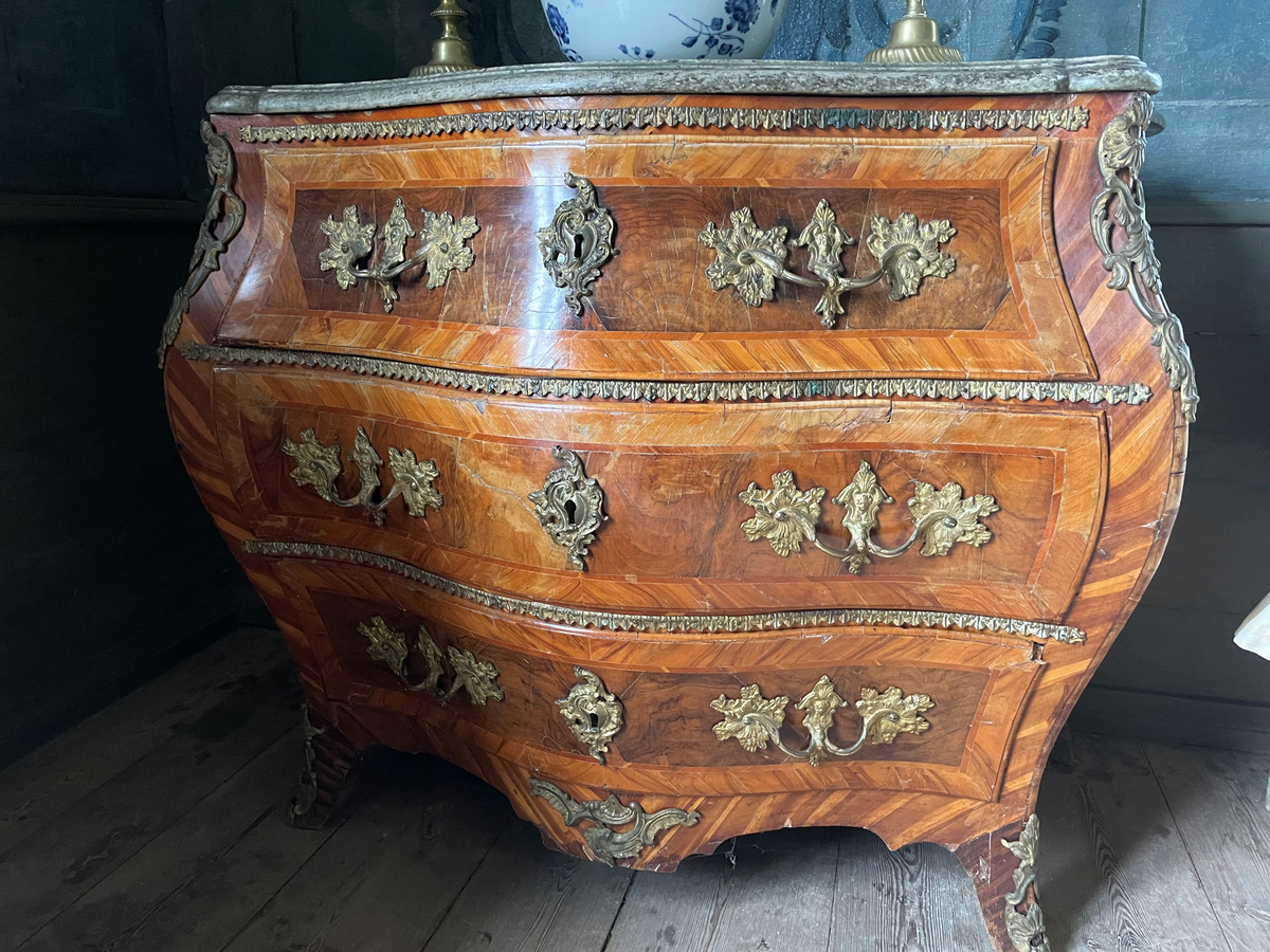 Byrå i rokoko med skiva av kolgårdsmarmor. Byrån har fanér av bland annat äppel- eller plommonträ, alm och valnötsrot. Beslag av brons, ursprungligen brännförgyllda. 

2024 gjordes en konservering av faner och rengöring av beslag.