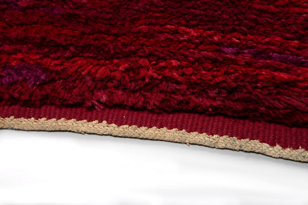 Stort gulvteppe/rye i ull i ulike rødnyanser og fiolett. Vevd teppe med floss på oversiden.