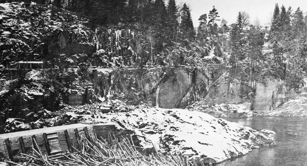 Fra Fossumfossen i den nedre delen av Glomma.  Fotografiet er tatt vinterstid, i en fase med et tynt snølag på marka og meget lav vannføring i elva.  Til venstre i bildeflata skimtes en slags dam eller terskel (skjerm), og nedenfor denne hadde det hopet seg opp fløtingstømmer som lå hulter til bulter.  Ellers preges landskapet på bildet av forholdsvis steile bergskrenter mot elveløpet med gran- og furuskog lengre oppe på land.  På ei utsprengt "hylle" i det nevnte berget later det til at det gikk en vegtrasé. 