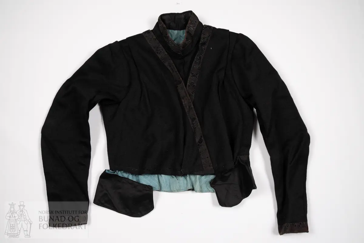 Kjoleliv av svart, kypertvevd ull. 
Kanta med svarte ullbånd med innvevd svart mønster. 
Foret er av bomullslerret med blå bunn, og smale hvite striper.
Midt foran lukkes dette stoffet som en bluse, med hekter. Øverst er det påsydd en skrå kant av det svarte stoffet, dette lukkes med trykknapper.
Livet har høy hals, kanta med bånd, denne lukkes med hekter. Over dette legges nok et lag med det svarte ullstoffet, dette lukkes midt foran med en trykknapp. Og gir en v-ringing som gjør halsen og deler av det svarte stoffet under synlig. 
Livet har et legg på hver skulder, som er med på å gi volum til brystpartiet.
Tosømsermer med litt puff, kanta nederst med bånd.
Nederst er det festa en kappe av svart bomullssateng, denne samler vidden i livet.
Midt bak er det festa tre hekter øverst på denne kappa. 
Kappa er delvis spretta av, den henger ikke lenger på livet foran. 
Nederst på ermene er livet fora med den samme svarte bomullsatengen som kappen er sydd av.
Maskinsydd med noen håndsydde detaljer.