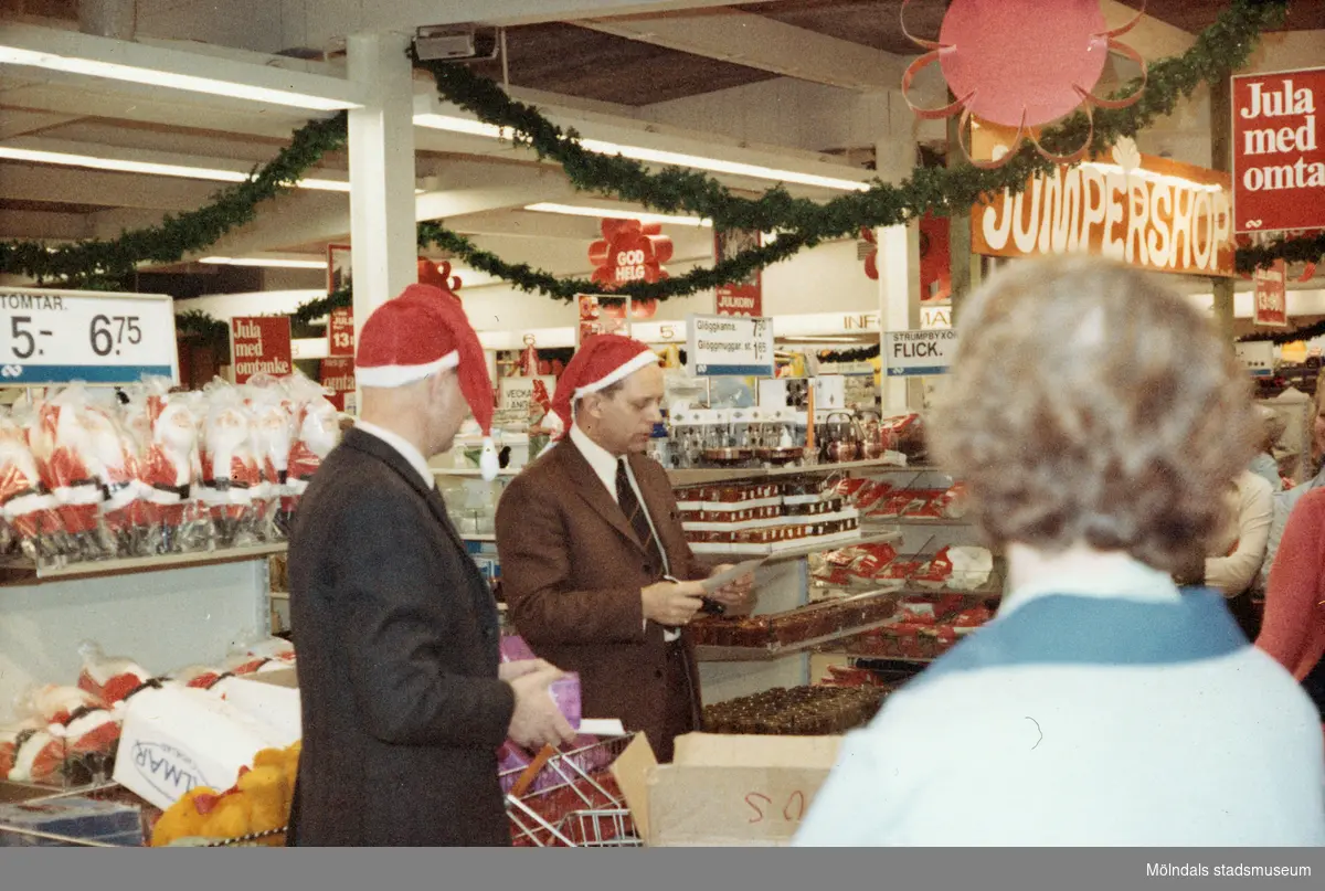 Kjell och Thore har på sig julmössor och delar ut julklappar till kunderna, Domus vid Nya torget julafton 1968. I bakgrunden ses en skylt som hänger i taket med texten "JUMPERSHOP". Gröna gran-girlanger hänger i taket som dekoration. 
Det är samma personer som på foto 2024_1184.
