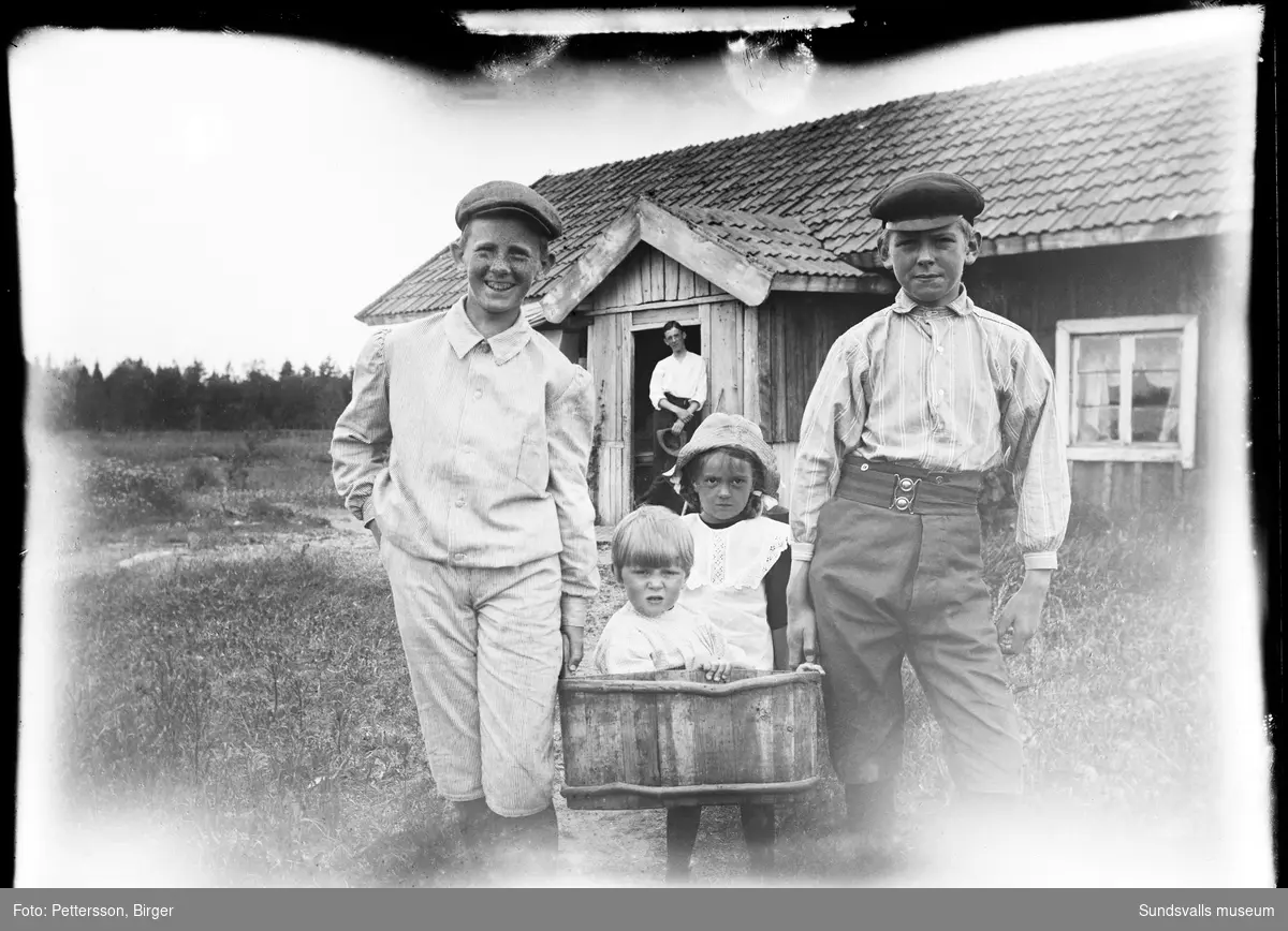 Gruppbild med två små och två större barn framför ett mindre trähus/stuga, sannolikt på Brämön. En man står i stugans dörröppning.