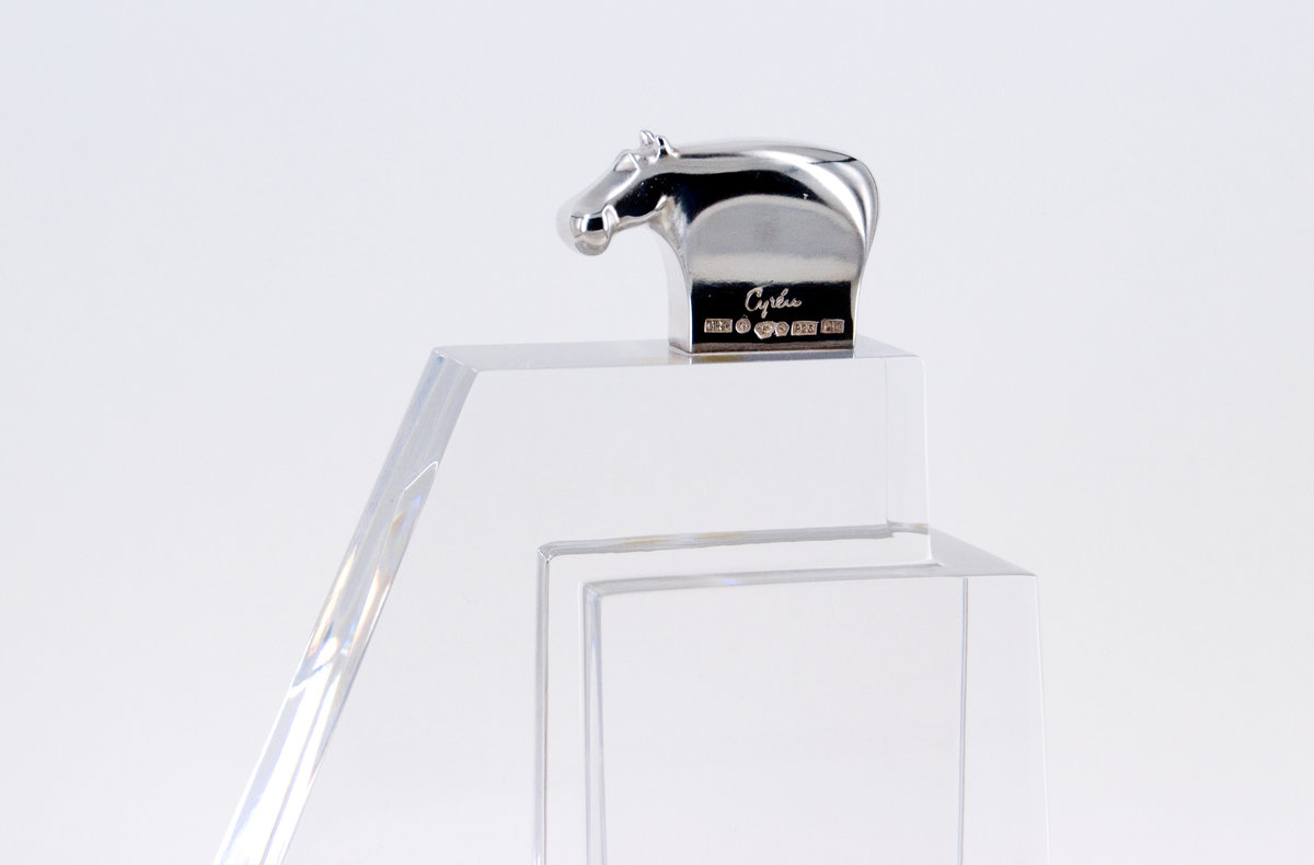 Skulptur, "Litet tempel", i silver och glas, av Gunnar Cyrén, 1989. Högt glasfundament med T-formad bas. Avsmalnande, delvis trappstegsformad, uppåt. Krönt av liten flodhäst i silver.
