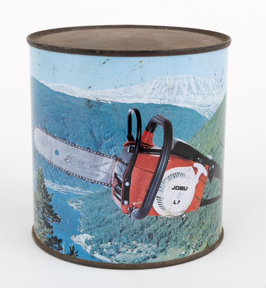 En sylindrisk boks, blikkboks, reklamemateriell, fra motorsagprodusenten / salgsbedriften Jo-Bu i Drøbak. Boksen har påtrykt firmaets logo og et bilde av motorsaga Jo-Bu (JOBU) L7. Saga svever over et norsk fjordlandskap med skogkledte fjellsider i forgrunnen og snødekte fjelltopper i bakgrunnen. Logoen som er påtrykt blikkboksen består av navnet JO BU med et grantre mellom JO og BU. Navnet omkranses av et sagkjede. Under "sagkjedet" står teksten: "OSLO-Norway".

Boksen har ukjent innhold, men en hører en raslelyd når den ristes.