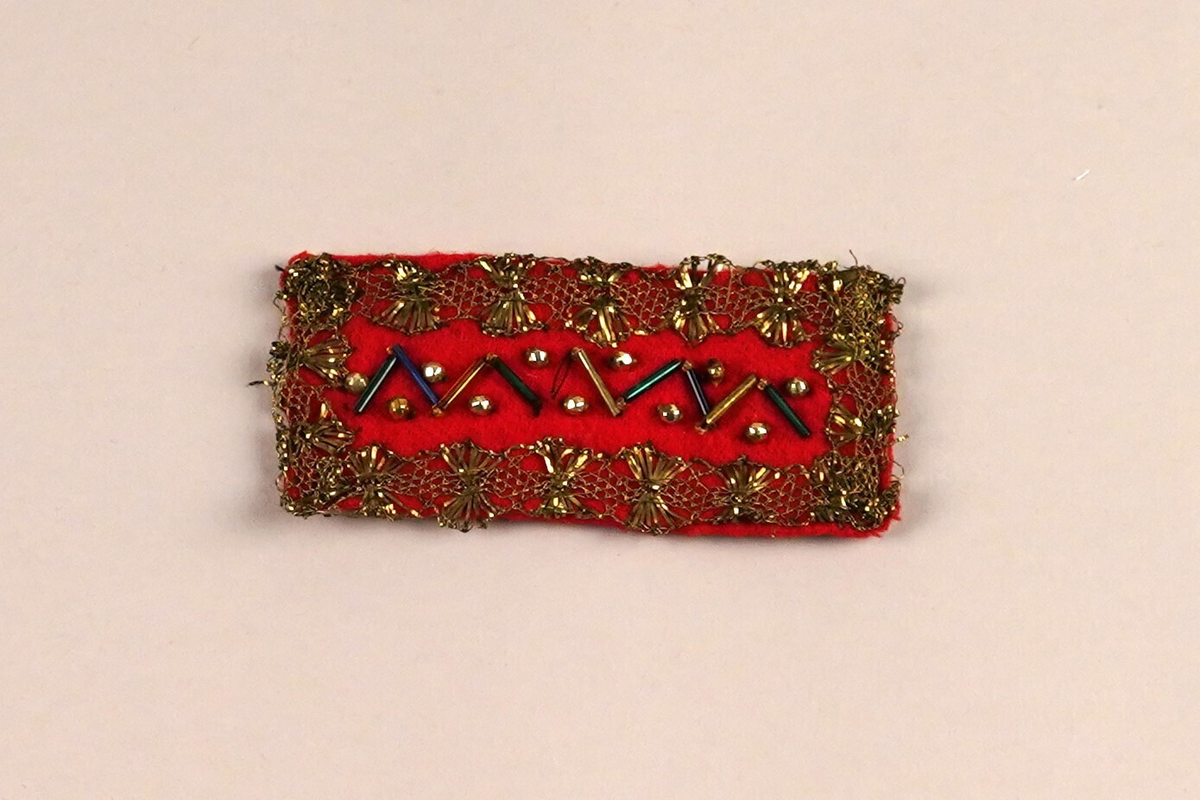 Band. Til å ha over skøyten bak på pannebora (panneband NFM.1995-00180). Ull stoff påsydd gull metallband, perler i midtfeltet. 