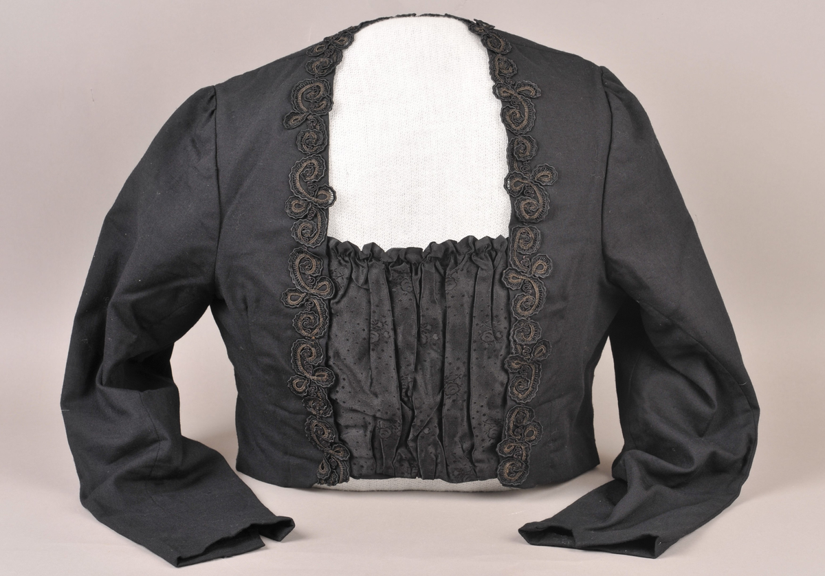 Svart stastrøye med smekke i damask-vevd silke. På sida av smekka er det sauma på smale kaupeband som er lagt i rosemønster. Fòra med svart bomullsstoff, med innvendig kneppa liv. Armane er fòra med svart silke.