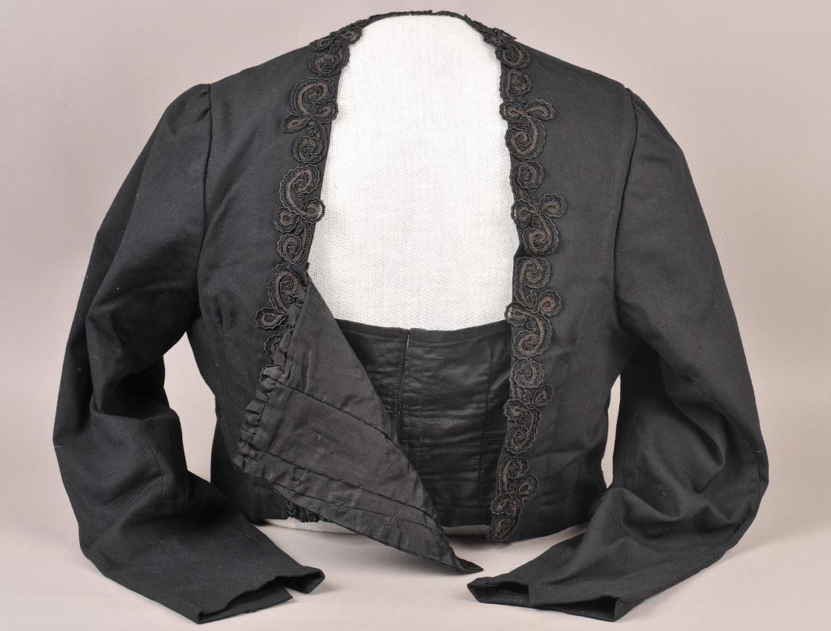 Svart stastrøye med smekke i damask-vevd silke. På sida av smekka er det sauma på smale kaupeband som er lagt i rosemønster. Fòra med svart bomullsstoff, med innvendig kneppa liv. Armane er fòra med svart silke.