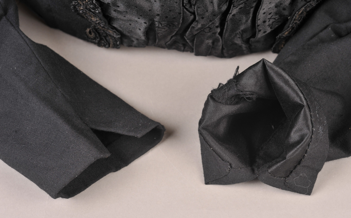 Svart stastrøye med smekke i damask-vevd silke. På sida av smekka er det sauma på smale kaupeband som er lagt i rosemønster. Fòra med svart bomullsstoff, med innvendig kneppa liv. Armane er fòra med svart silke.