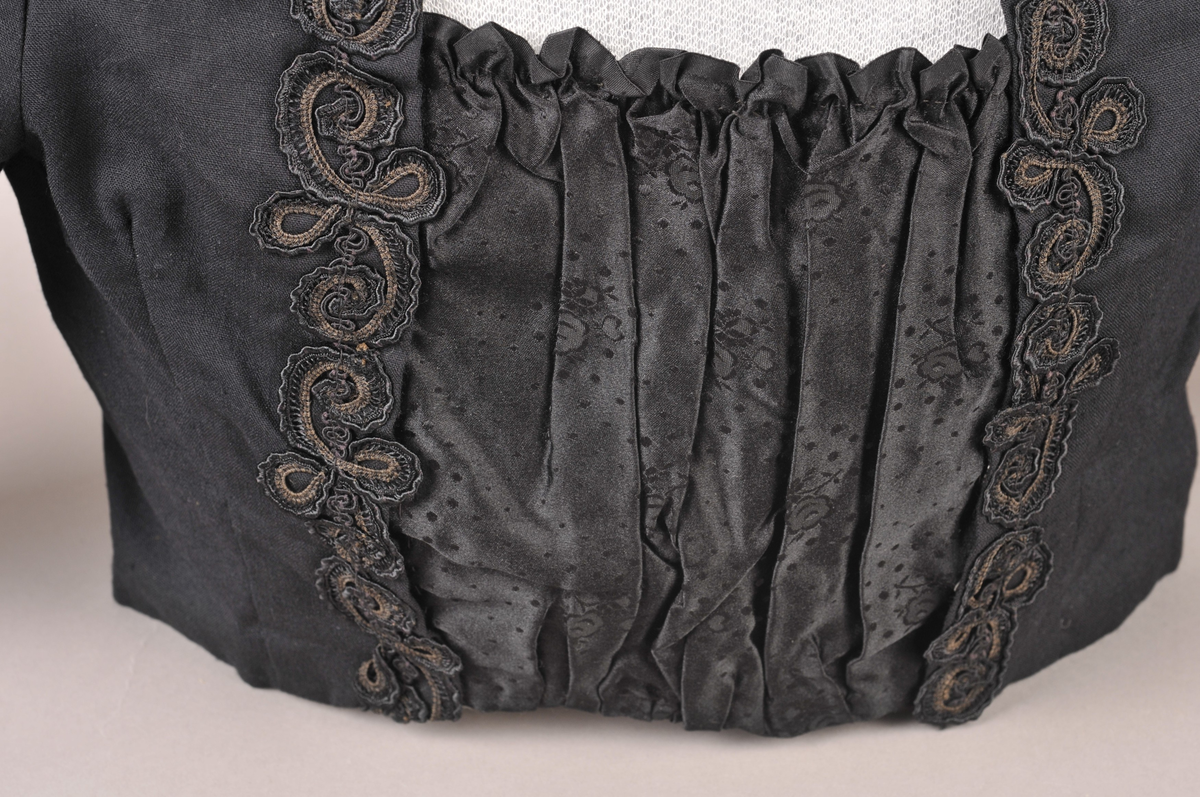 Svart stastrøye med smekke i damask-vevd silke. På sida av smekka er det sauma på smale kaupeband som er lagt i rosemønster. Fòra med svart bomullsstoff, med innvendig kneppa liv. Armane er fòra med svart silke.