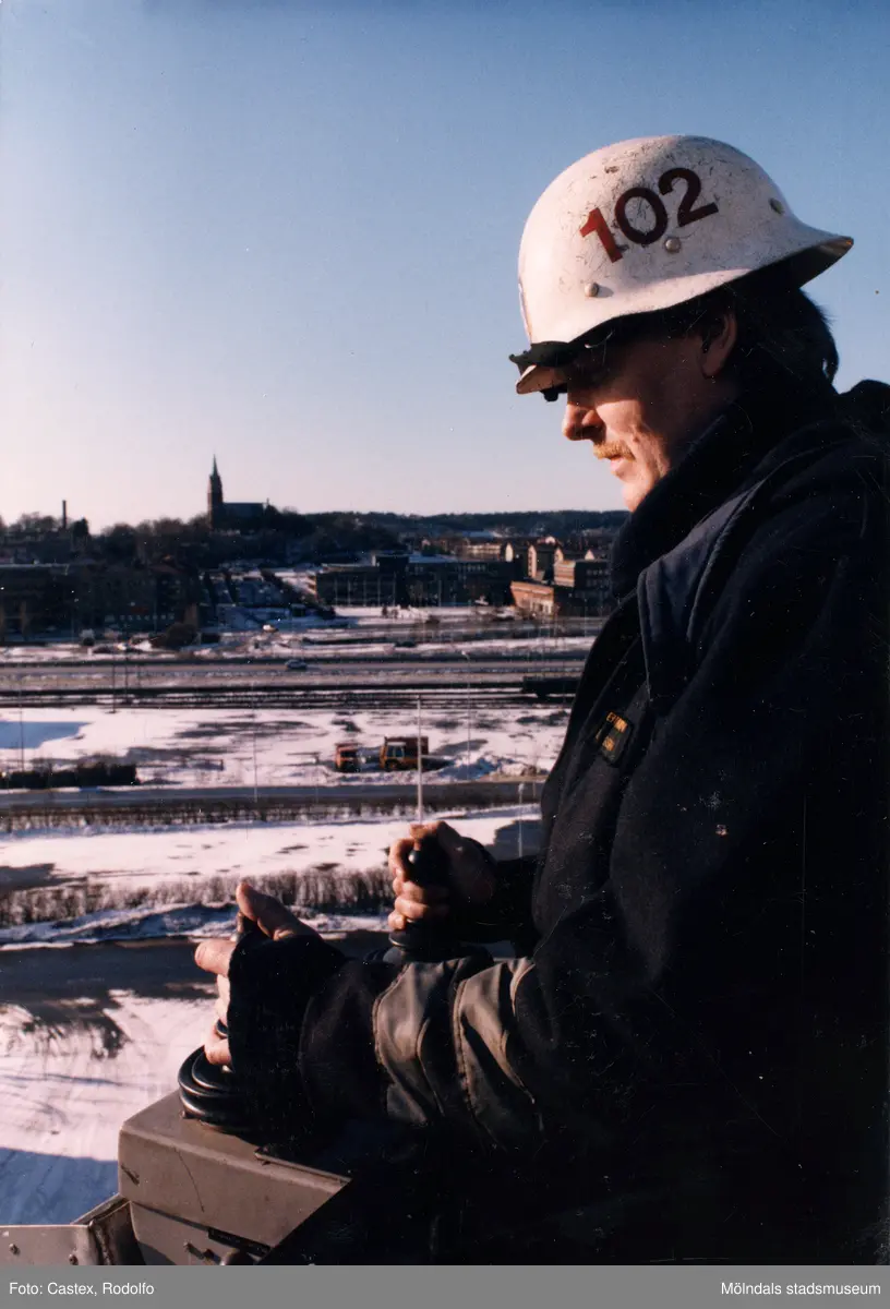 Mölndals brandstation i Trädgården, Mölndal, på 1980-talet. En brandman manövrerar brandbilens lift med hjälp av spakar. I bakgrunden ses Fässbergs kyrka, stadshuset och Folkets hus.