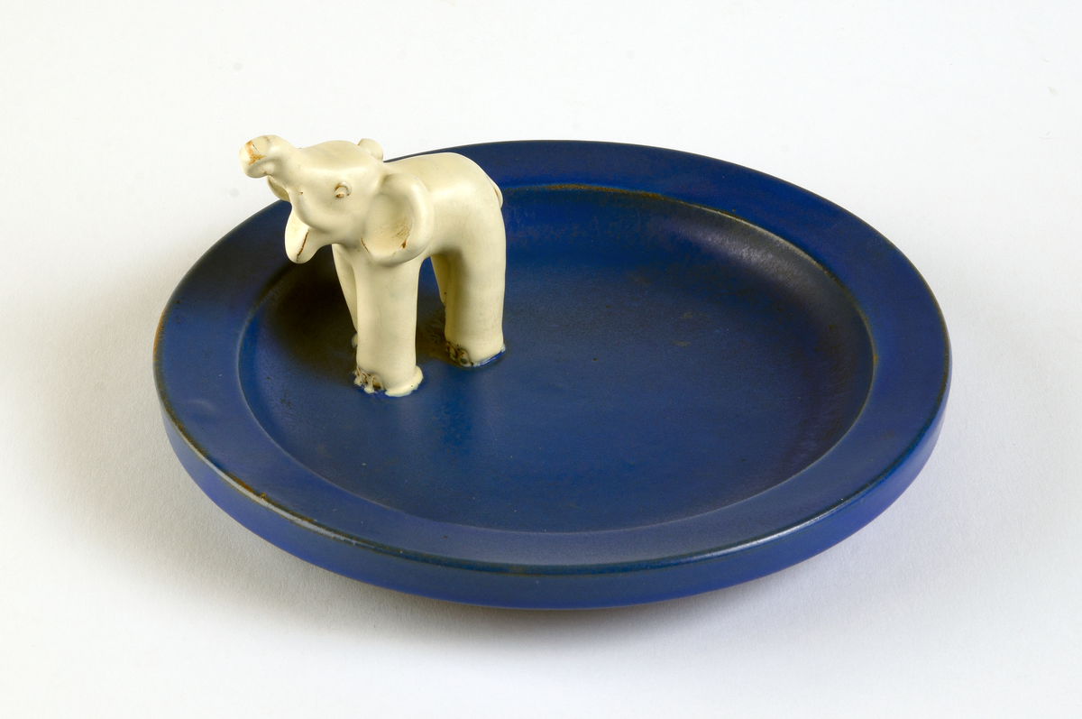 Runt askfat av lergods med halvmatt glasyr i blått, vid kanten  en vitglaserade modellerad elefant. Fatet gjutet. I botten stämplat: EKEBY 46.