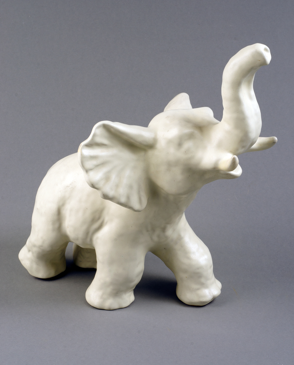 Prydnadsföremål i form av elefant med höjd snabel, av keramik med halvblank vit glasyr, gjuten.

