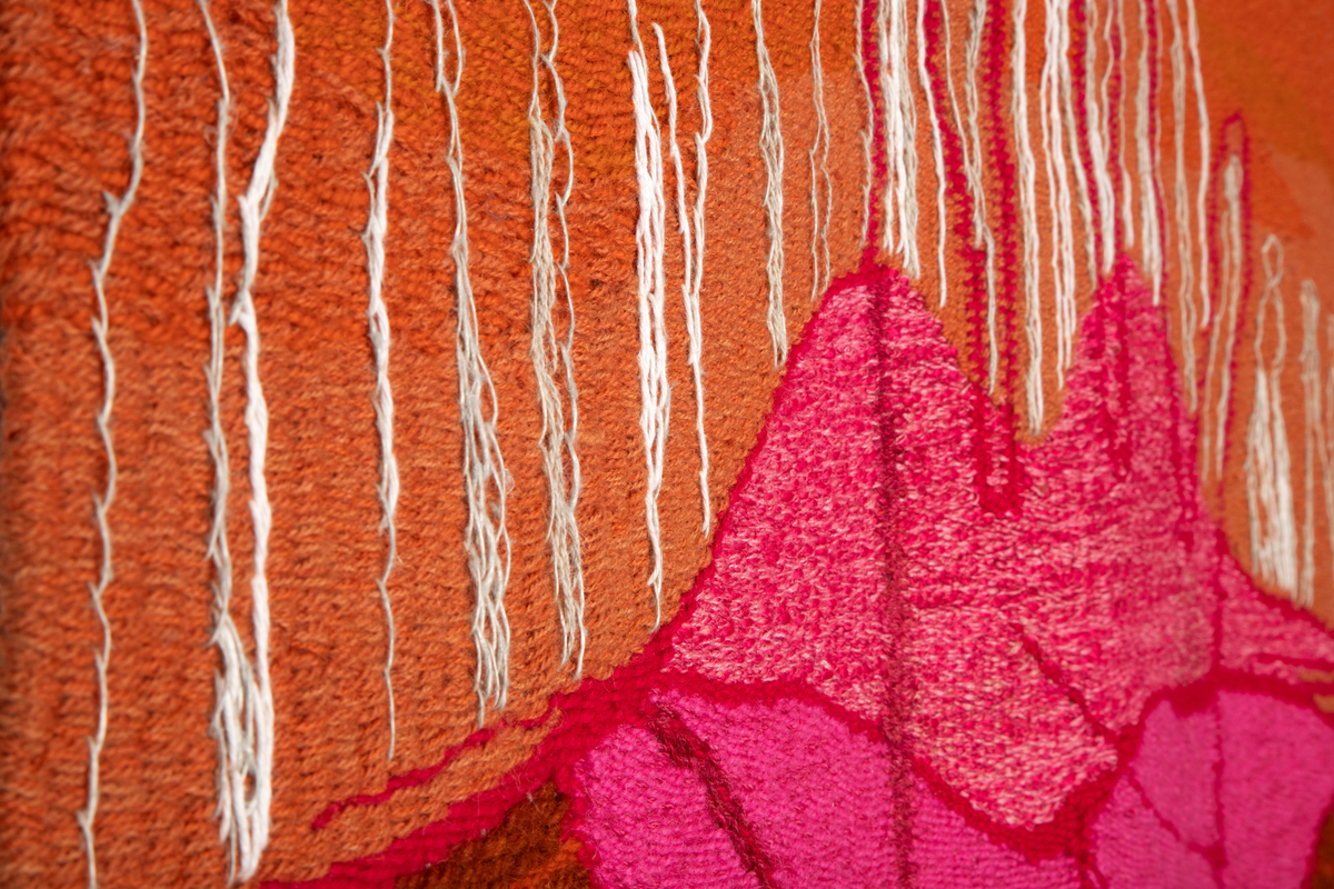 Håndvevd billedvev i tilnærmet kvadratisk format. Landskapslignende motiv i ulike nyanser av brunt, rødt og oransje med konturene av en stor hånd over rosa lepper. Hvitbroderte mennesker står/går i landskapet. Nederst i midten det internasjonale peace-merket.