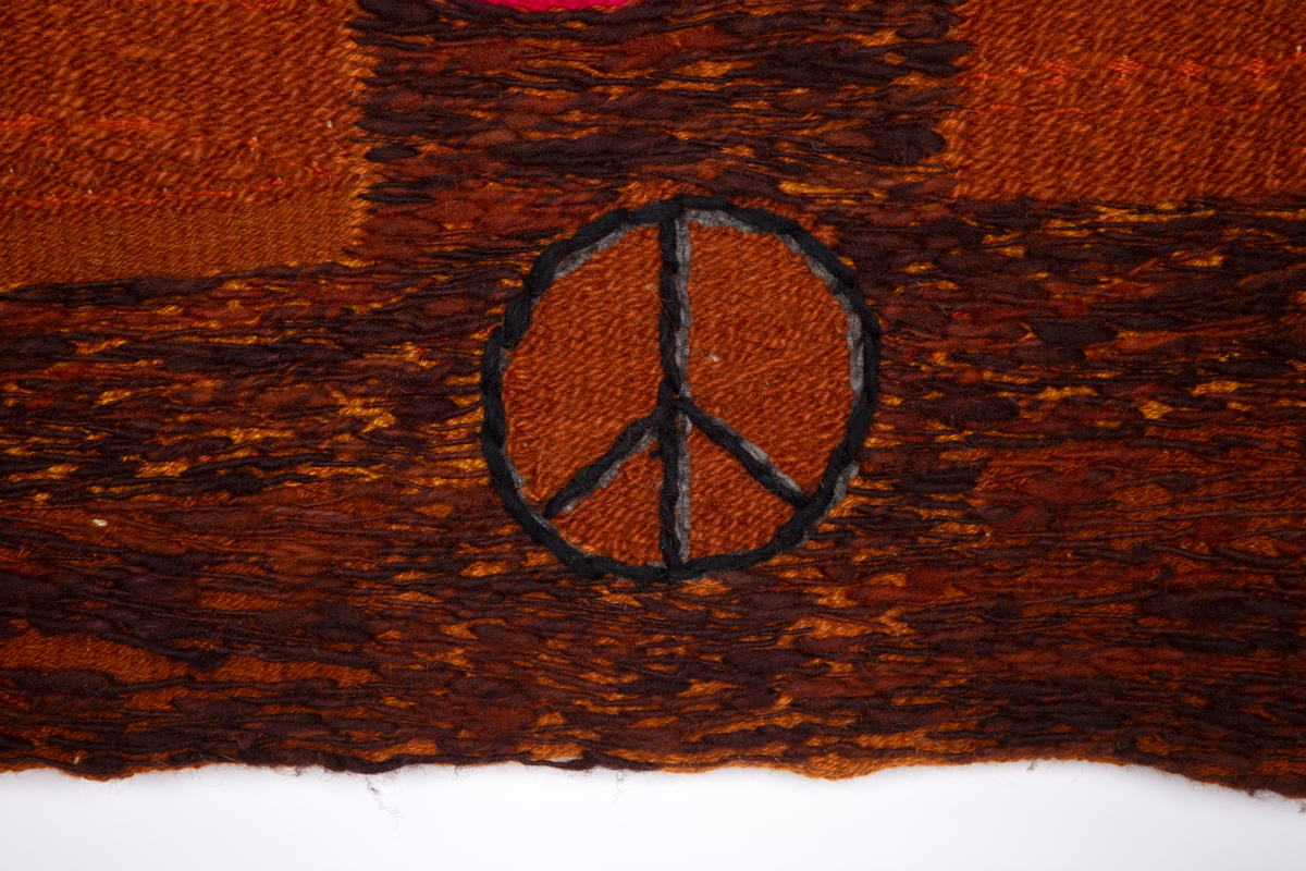 Håndvevd billedvev i tilnærmet kvadratisk format. Landskapslignende motiv i ulike nyanser av brunt, rødt og oransje med konturene av en stor hånd over rosa lepper. Hvitbroderte mennesker står/går i landskapet. Nederst i midten det internasjonale peace-merket.