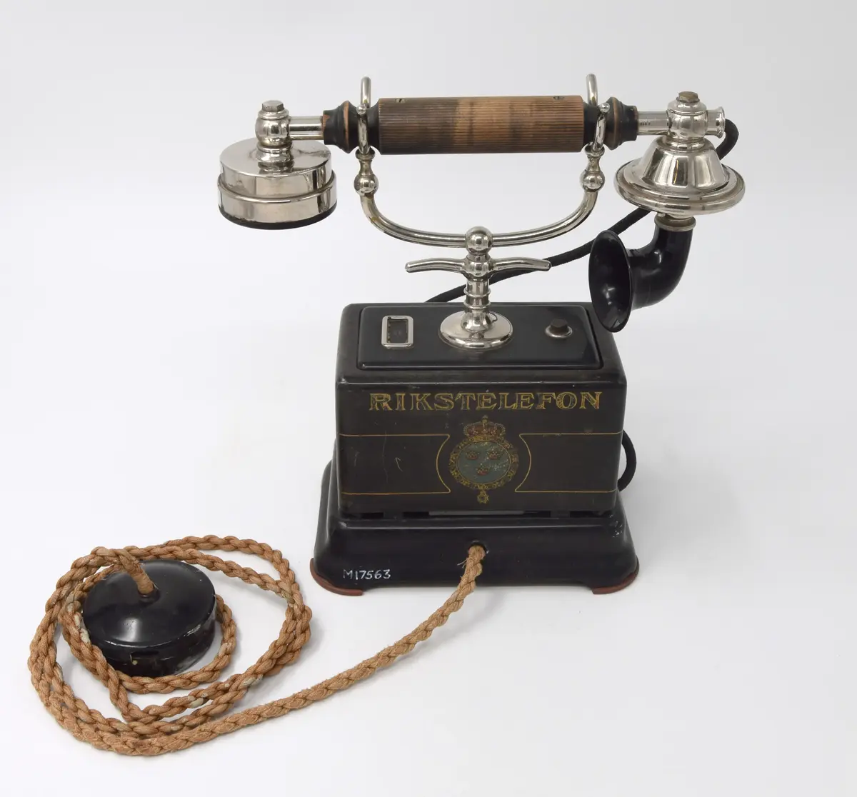 Telefonapparat, svart. Märkt: Rikstelefon och svenska lilla riksvapnet. Saknar fingerskiva. Klyka av vitmetall med grepp att bära telefonen i. Mikrotelefon av vitmetall med handtagskavel av svartmålat trä. Mikrofontratt av svart bakelit. Vid apparaten är en rund svart dosa, diam. 6,5 cm, med en grov brun sladd, L. 144 cm. 