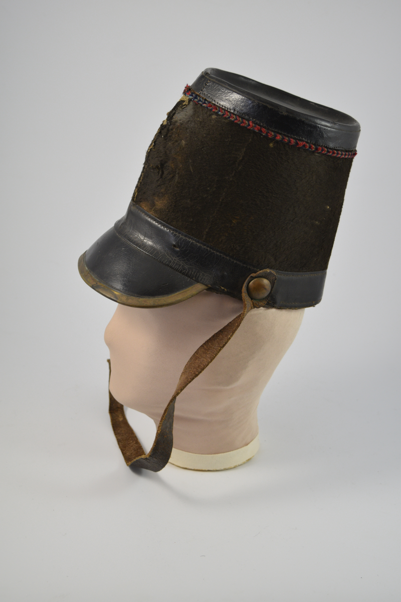 Sylinderformet hatt med brem. Bremmen avsluttes med en smal messingkant. Pullen er dekket med pelsliknende tekstil. En bred stripe av skinn øverst og nederst på pullen samt bremmen og toppen av hatten er dekket av skinn. Dekorativt bånd i rødt, blått og gull like under skinnkanten øverst på pullen.
