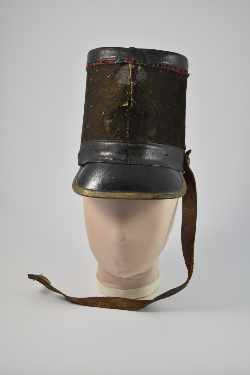 Sylinderformet hatt med brem. Bremmen avsluttes med en smal messingkant. Pullen er dekket med pelsliknende tekstil. En bred stripe av skinn øverst og nederst på pullen samt bremmen og toppen av hatten er dekket av skinn. Dekorativt bånd i rødt, blått og gull like under skinnkanten øverst på pullen.