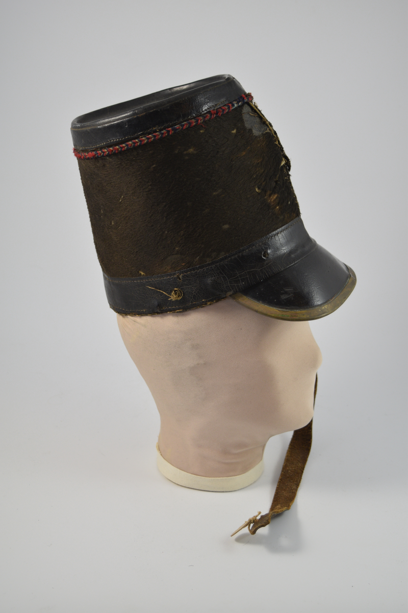 Sylinderformet hatt med brem. Bremmen avsluttes med en smal messingkant. Pullen er dekket med pelsliknende tekstil. En bred stripe av skinn øverst og nederst på pullen samt bremmen og toppen av hatten er dekket av skinn. Dekorativt bånd i rødt, blått og gull like under skinnkanten øverst på pullen.