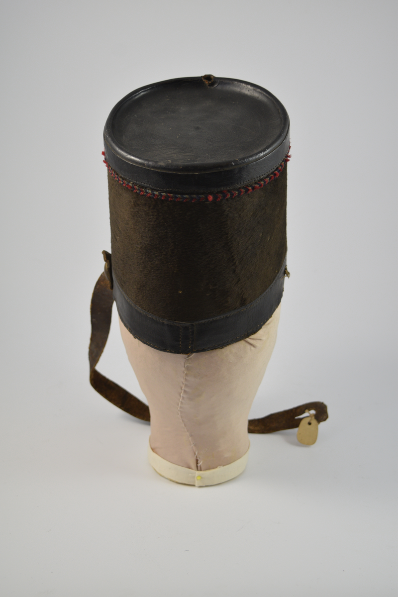 Sylinderformet hatt med brem. Bremmen avsluttes med en smal messingkant. Pullen er dekket med pelsliknende tekstil. En bred stripe av skinn øverst og nederst på pullen samt bremmen og toppen av hatten er dekket av skinn. Dekorativt bånd i rødt, blått og gull like under skinnkanten øverst på pullen.