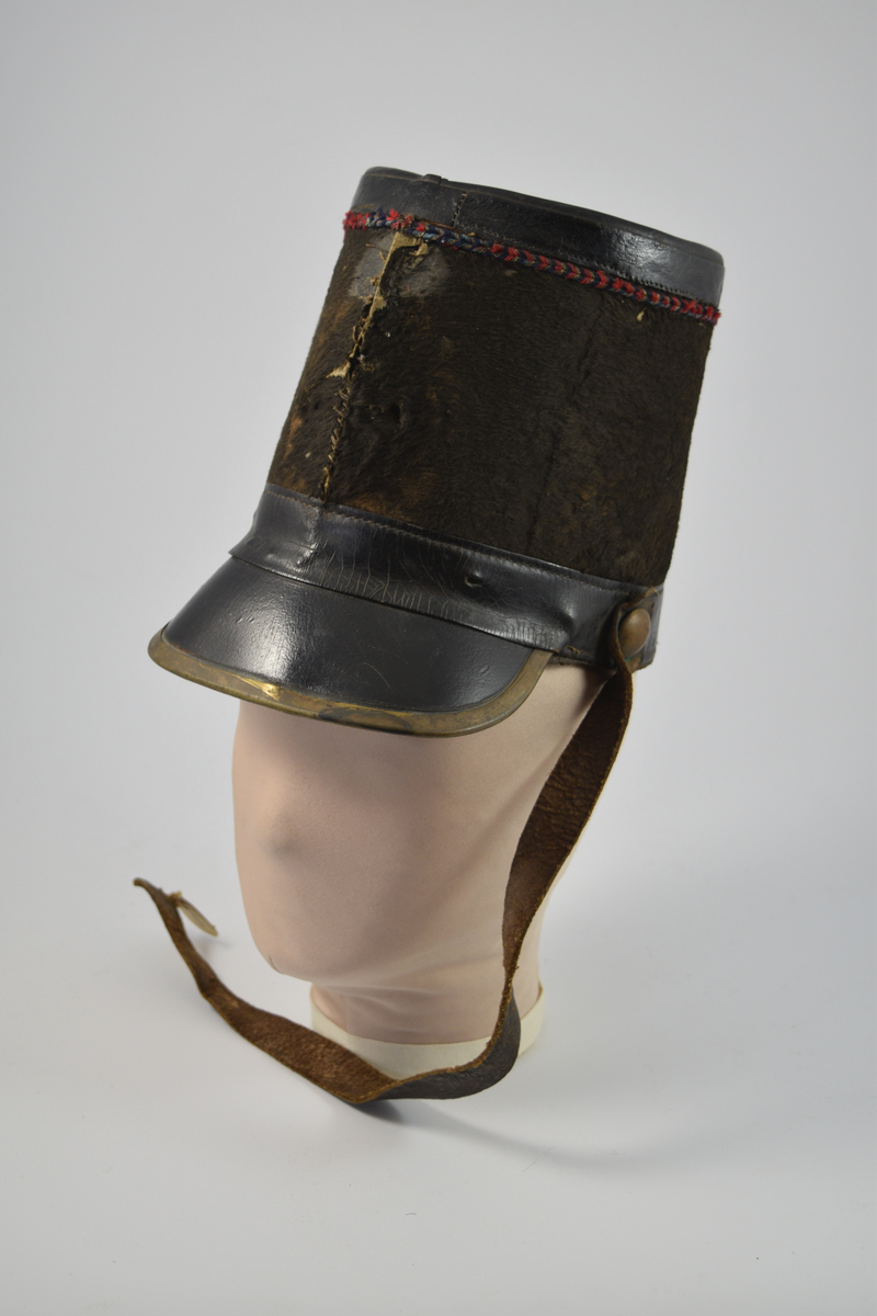 Sylinderformet hatt med brem. Bremmen avsluttes med en smal messingkant. Pullen er dekket med pelsliknende tekstil. En bred stripe av skinn øverst og nederst på pullen samt bremmen og toppen av hatten er dekket av skinn. Dekorativt bånd i rødt, blått og gull like under skinnkanten øverst på pullen.