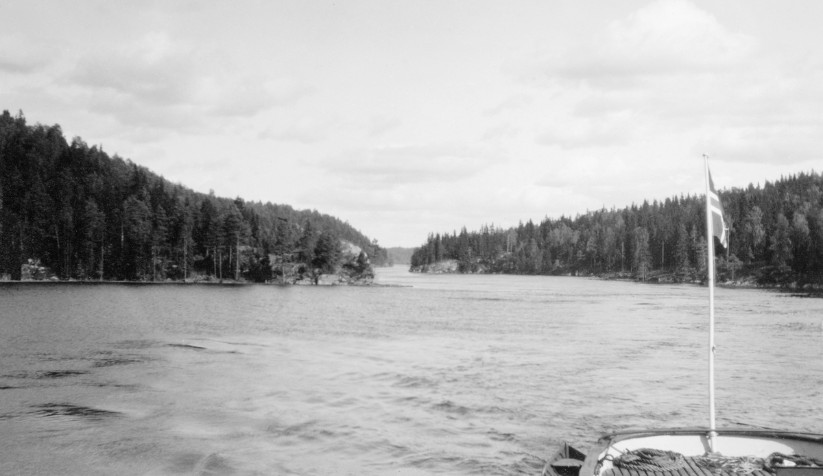 Parti fra Glommavassdraget, antakelig fra området ved Mørkfoss, altså like nedenfor Glommas utløp fra innsjøen Øyeren.  Fotografiet er tatt fra akterdekket på en fløtingsbåt med norsk flagg i akterstavnen.  Vi ser vassdraget omgitt av skogkledde berg og lier. 